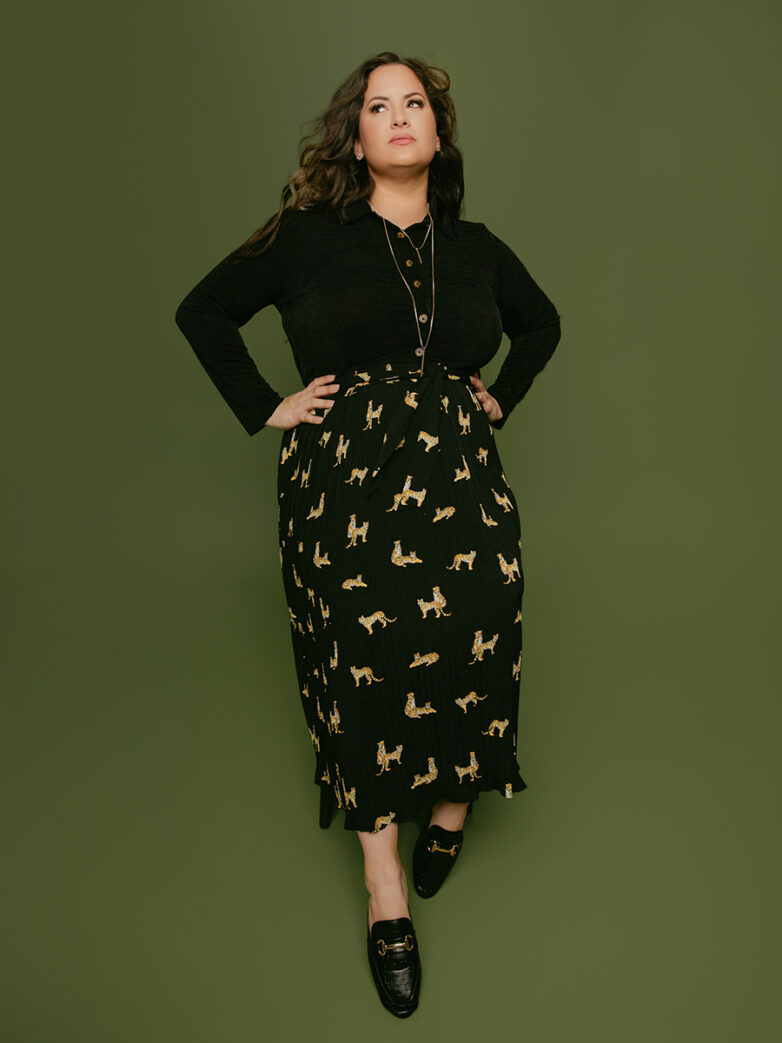 Woman in a black long-sleeve top and leopard-print midi skirt posing confidently against a green backdrop
