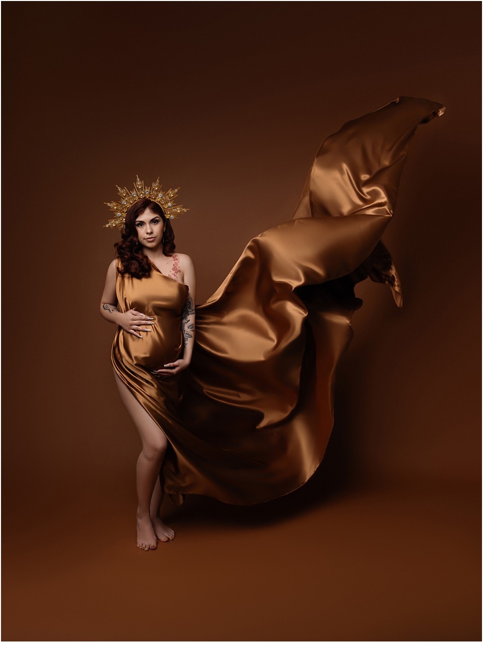 High-fashion maternity shoot with gold satin fabric flowing mid-air and a crown headpiece that resembles a goddess