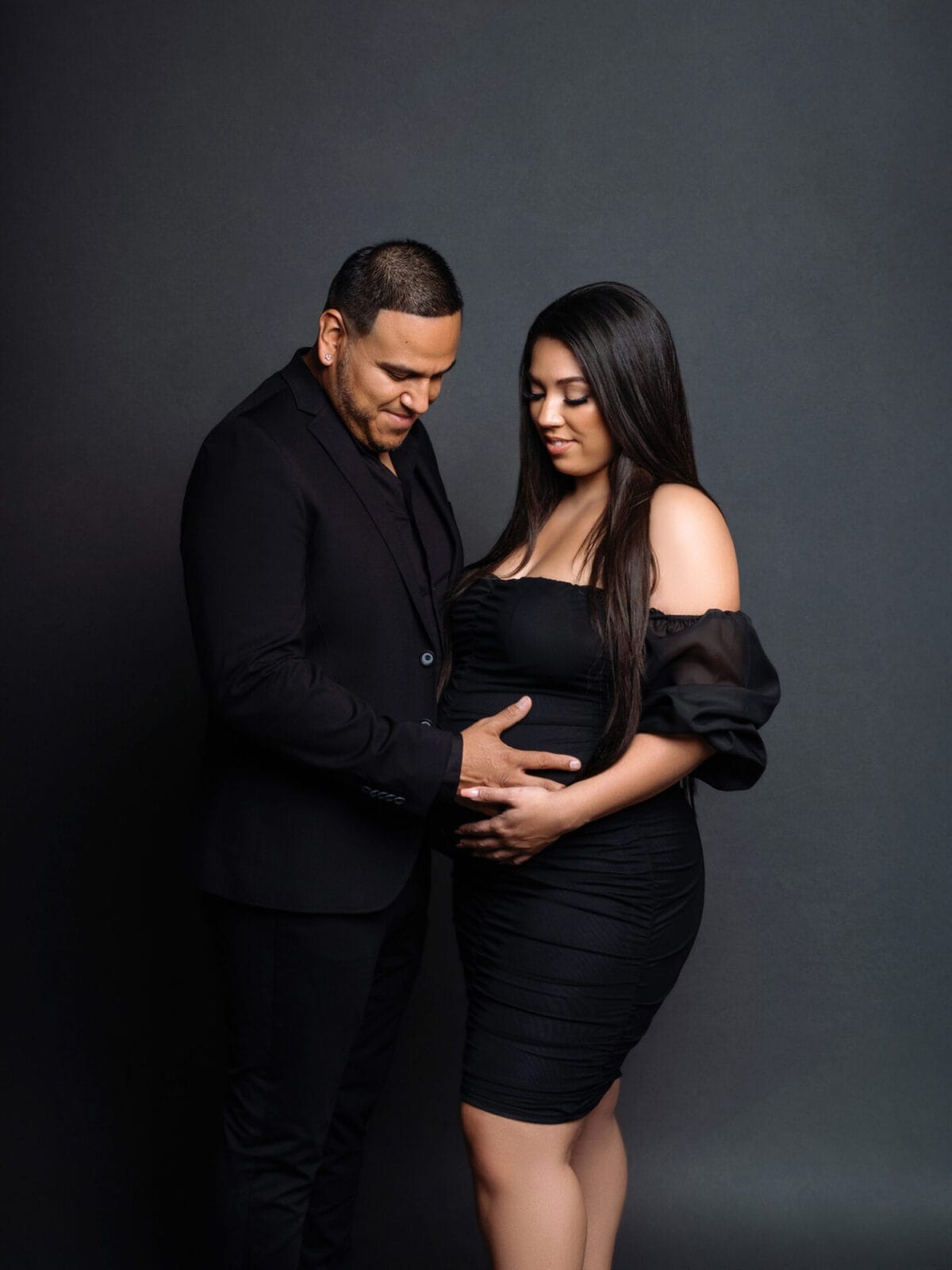 Elegant studio maternity portrait with couple dressed in all black, sharing a quiet moment while holding baby bump