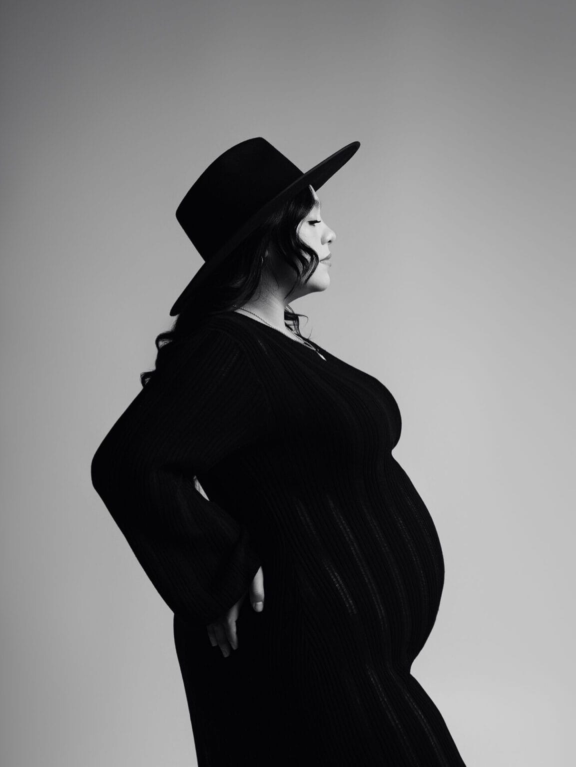Studio maternity portrait with mom wearing a wide-brim hat and form-fitting black dress, posed in profile.