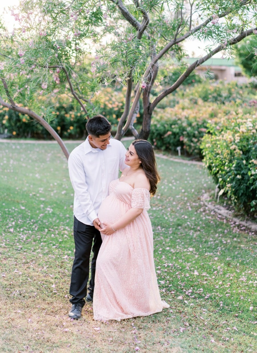 Rose-inspired outdoor maternity session with couple in a lush green garden under a blooming tree