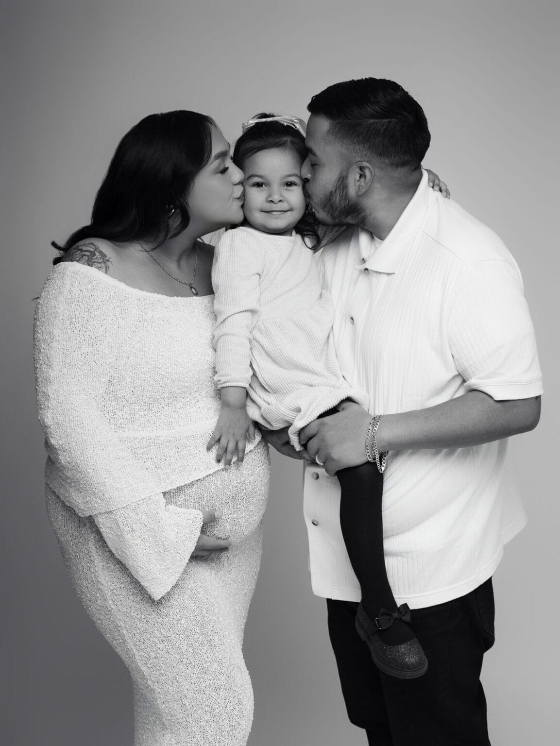 pregnant mom and dad kissing toddler on either side while mom is holding her pregnant belly, all dressed in light neutral tones.