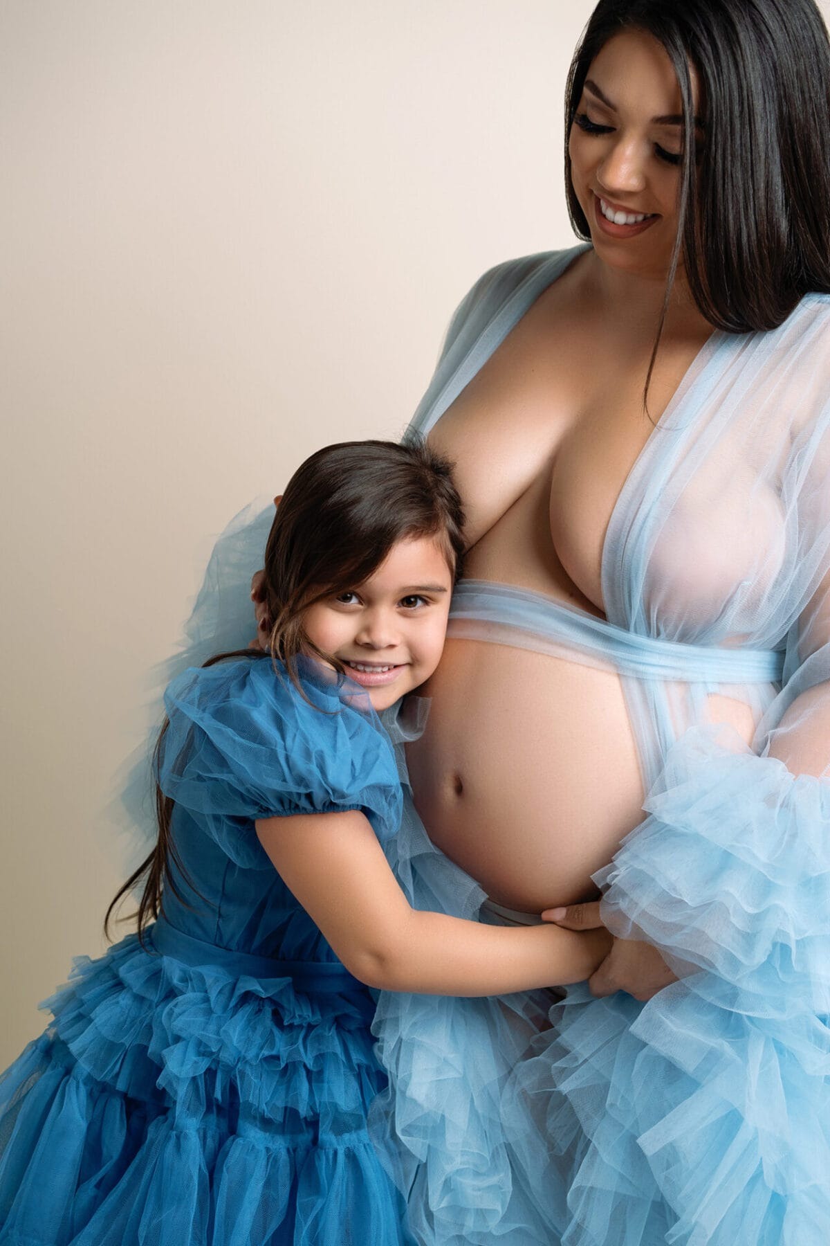 Sweet sibling moment during maternity session with little girl hugging mom’s baby bump, both in matching blue tulle dresses.
