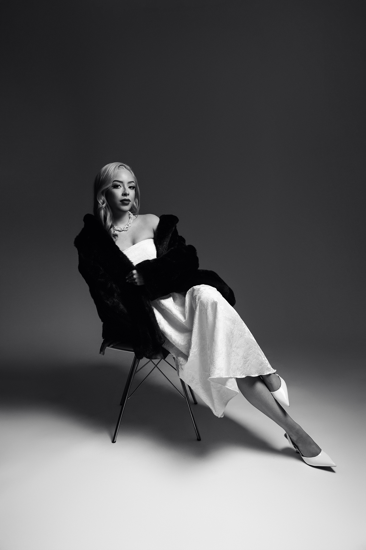 studio photo of a woman on a chair wearing a fur coat and glam dress with dramatic lighting