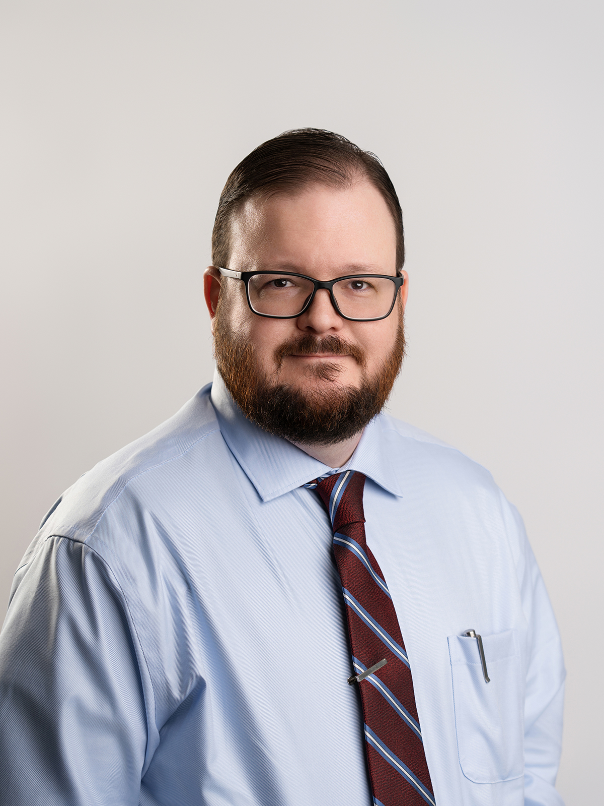 Professional headshot of a man in a light blue dress shirt, glasses, and striped tie