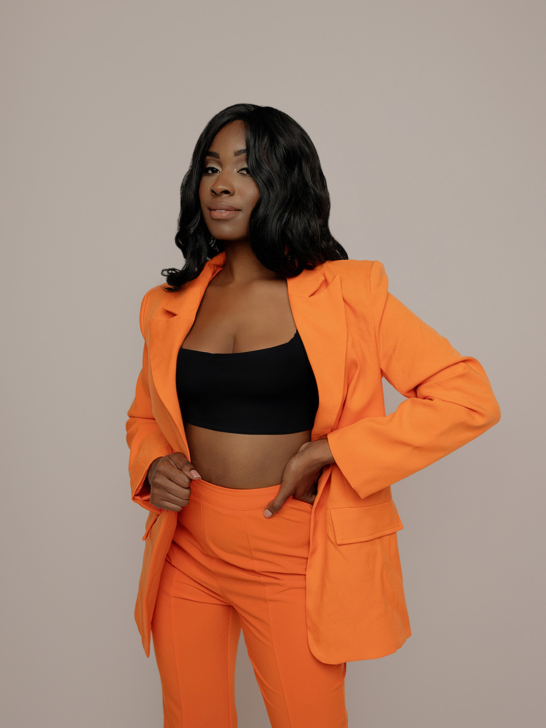 Bold studio portrait of a woman in an orange suit and black crop top on a beige backdrop