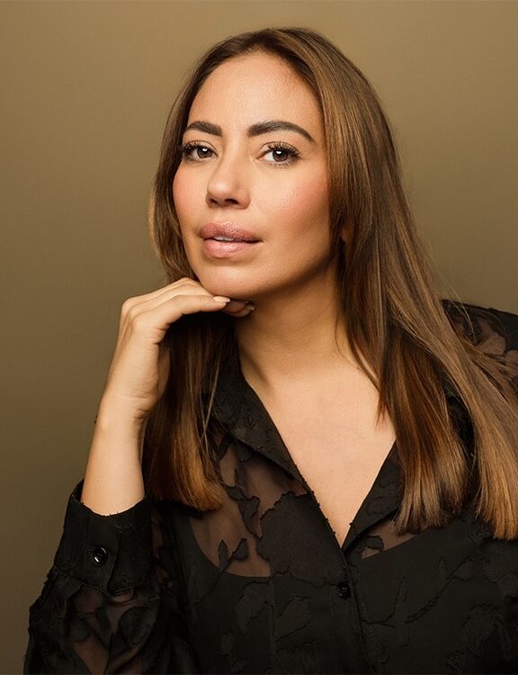 Confident studio headshot of a female actor with long brown hair, wearing a sheer black top on a tan background