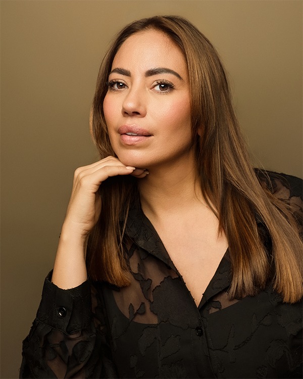 Confident studio headshot of a female actor with long brown hair, wearing a sheer black top on a tan background