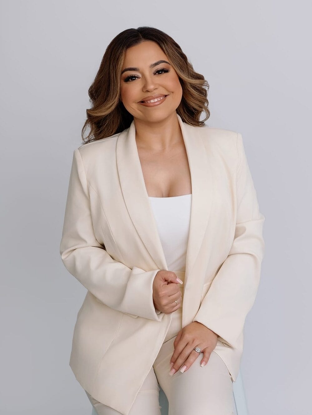 Elegant studio portrait of a smiling realtor woman in a cream suit seated on a light gray background