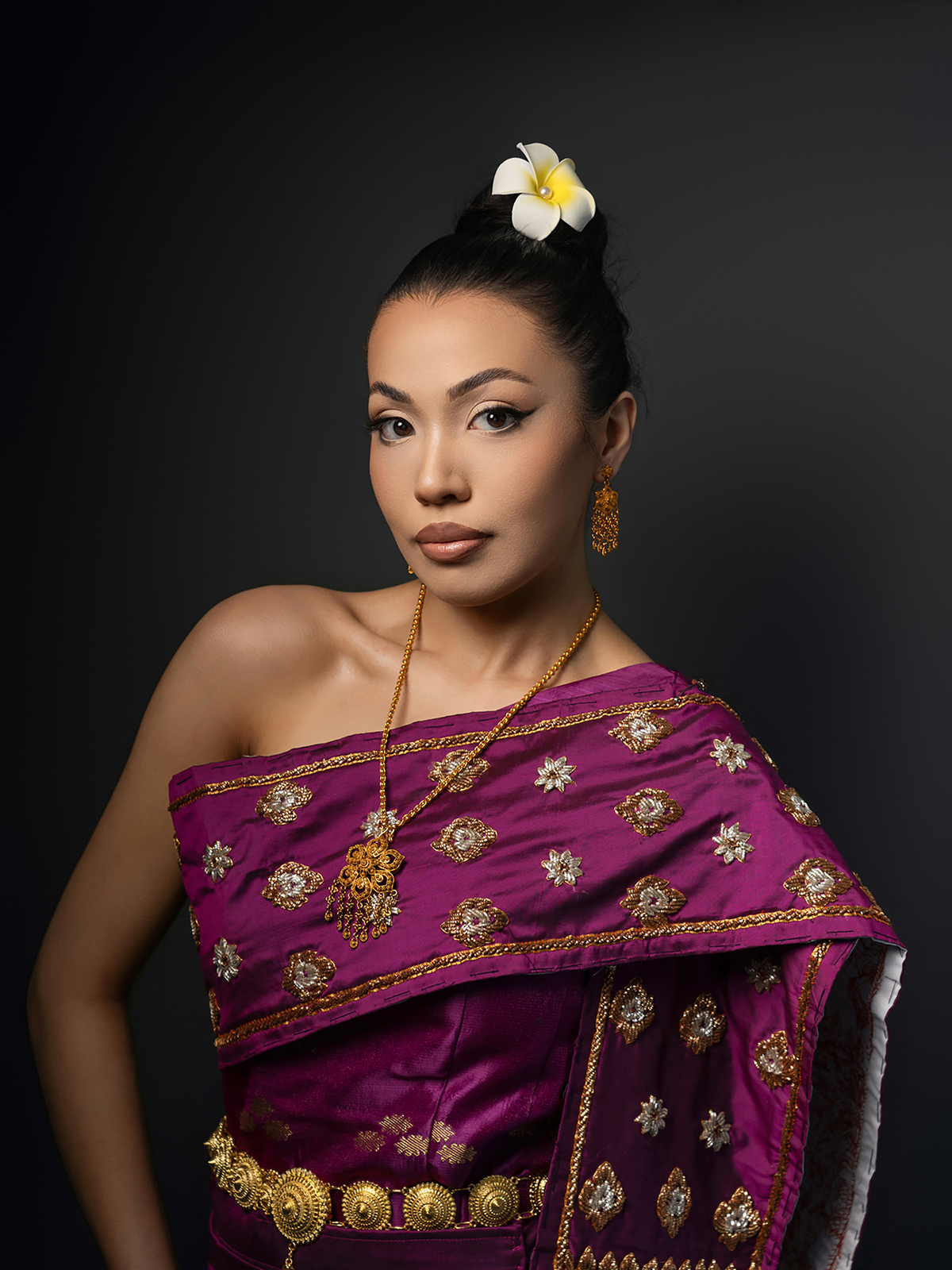 Studio pageant headshot of Miss Arizona contestant wearing traditional Thai-inspired magenta dress with gold jewelry, posed on dark backdrop
