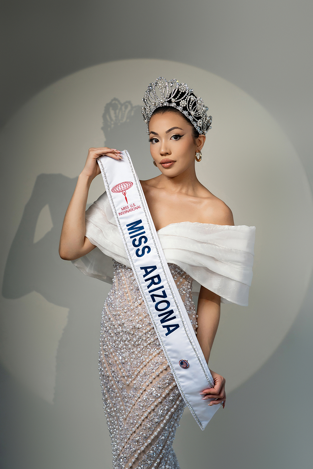 Close-up of Miss Arizona winner holding her Miss USA International sash and crown, wearing an off-shoulder pearl gown on gray backdrop