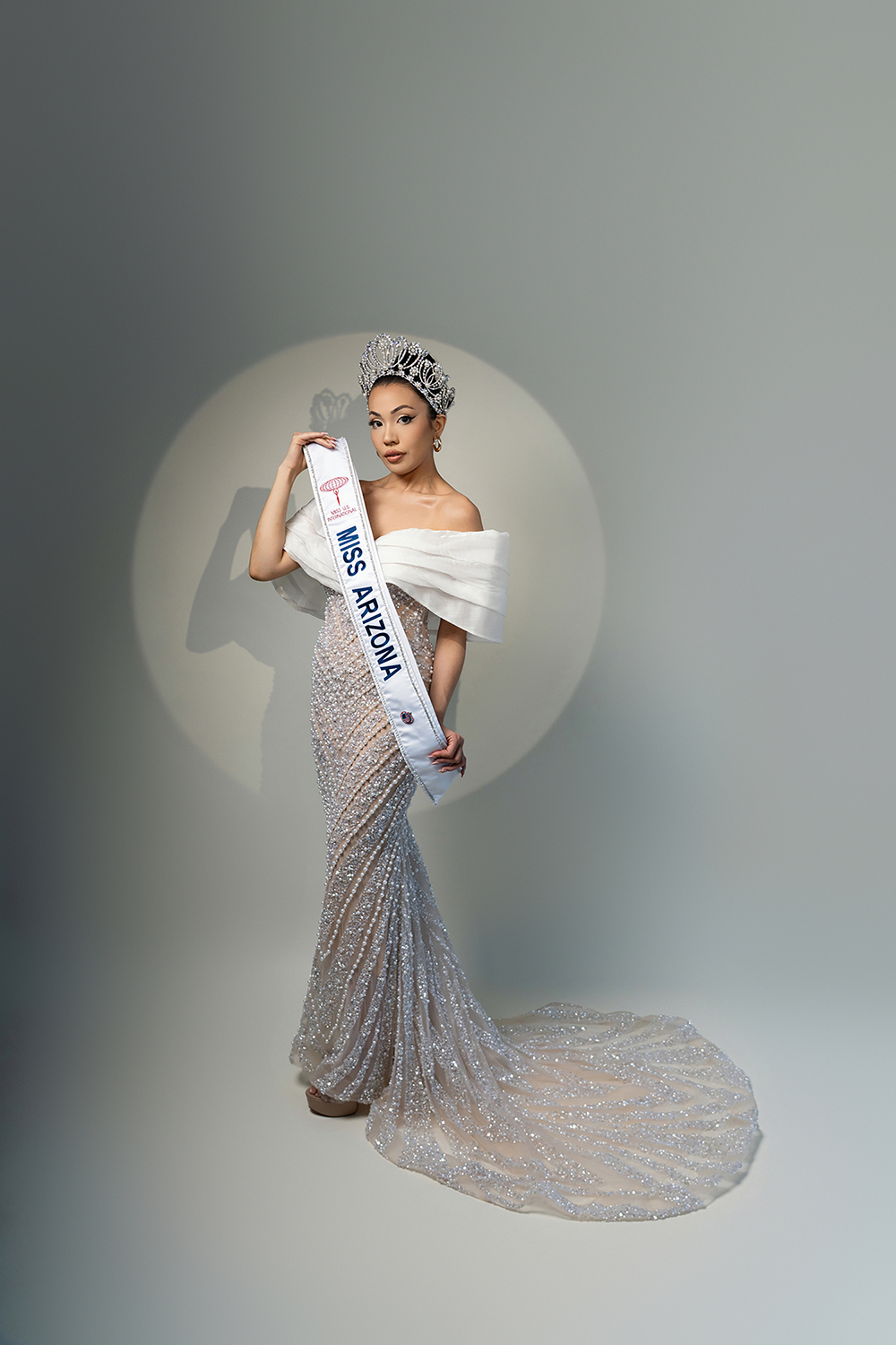 Full-body pageant portrait of Miss Arizona in sparkling pearl gown and crown, holding sash in studio lighting