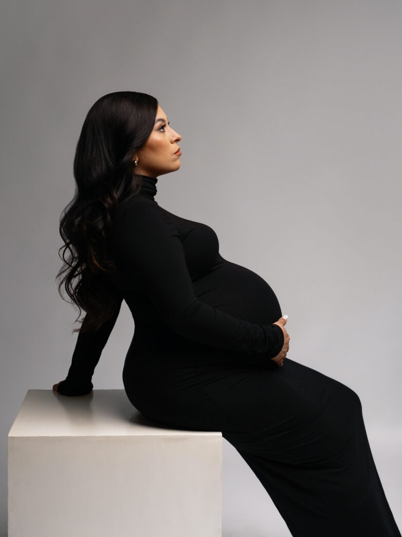 pregnant woman sitting on a posing box with a black maxi dress holding her dump in a studio session