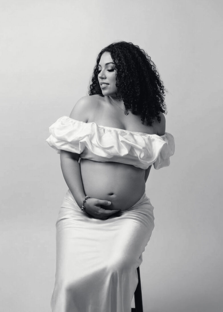 Black and white maternity portrait of an expectant mother wearing an off-shoulder white dress