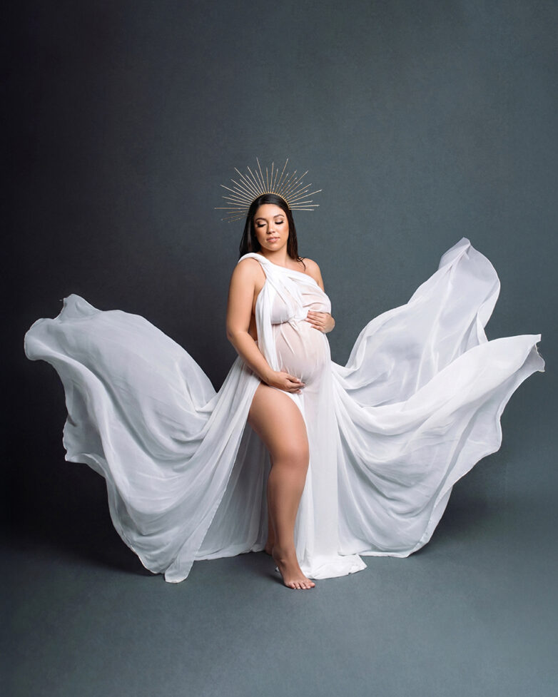 Pregnant woman in a flowing white fabric dress posing against a dark backdrop