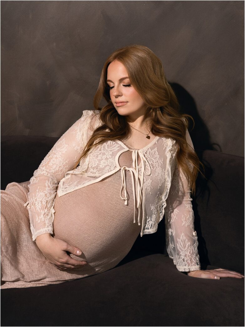 Expectant mother seated in a lace maternity robe cradling her baby bump