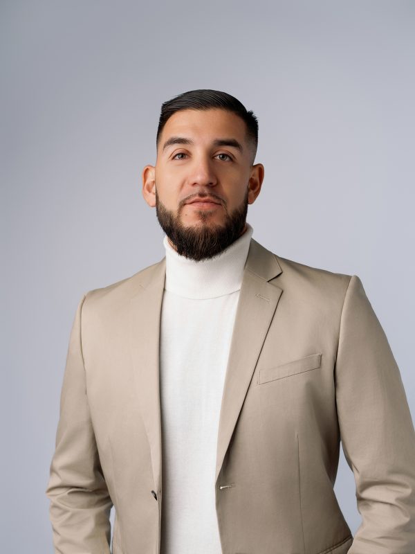 Man with a beard wearing a beige suit and white turtleneck, standing against a gray background